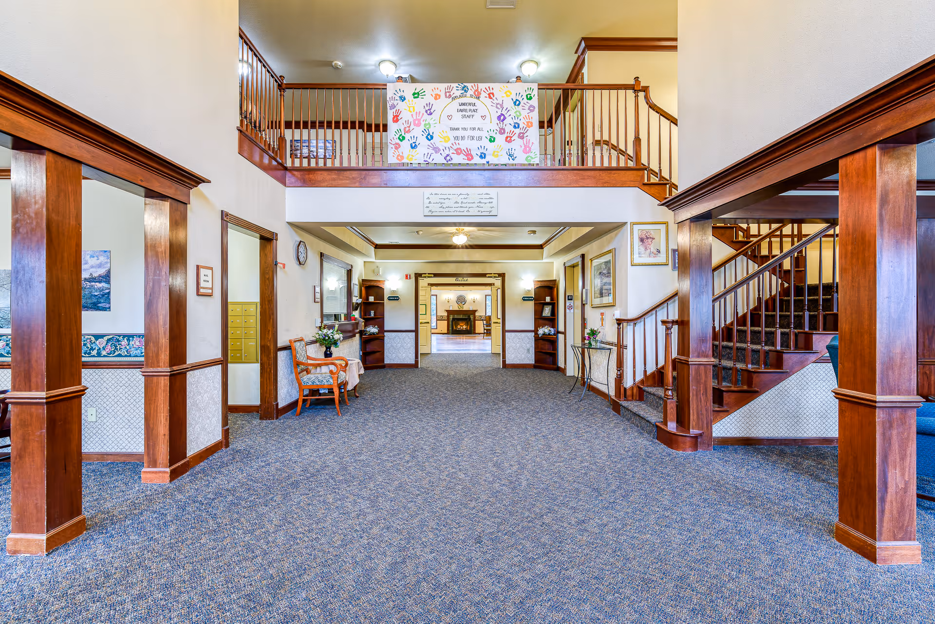 Interior view of a spacious assisted living facility lobby with carpeted floor, wooden pillars, and a staircase on the right. There is a balcony with a colorful handprint banner thanking the staff. The hallway leads to a room with a fireplace and seating area. Chairs and small tables with flowers are placed along the walls.