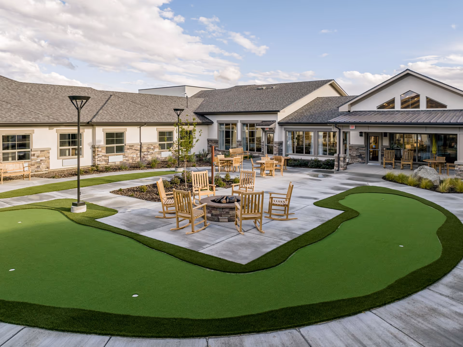 Outdoor courtyard area of Kiley Ranch Senior Living featuring a putting green, several wooden chairs arranged around a fire pit, additional seating areas with tables and chairs, and a building with large windows and stone accents under a partly cloudy sky.