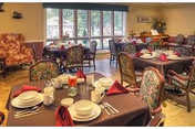 A bright dining room with multiple tables set with plates, cups and folded napkins, floral-upholstered chairs, and large windows overlooking greenery.