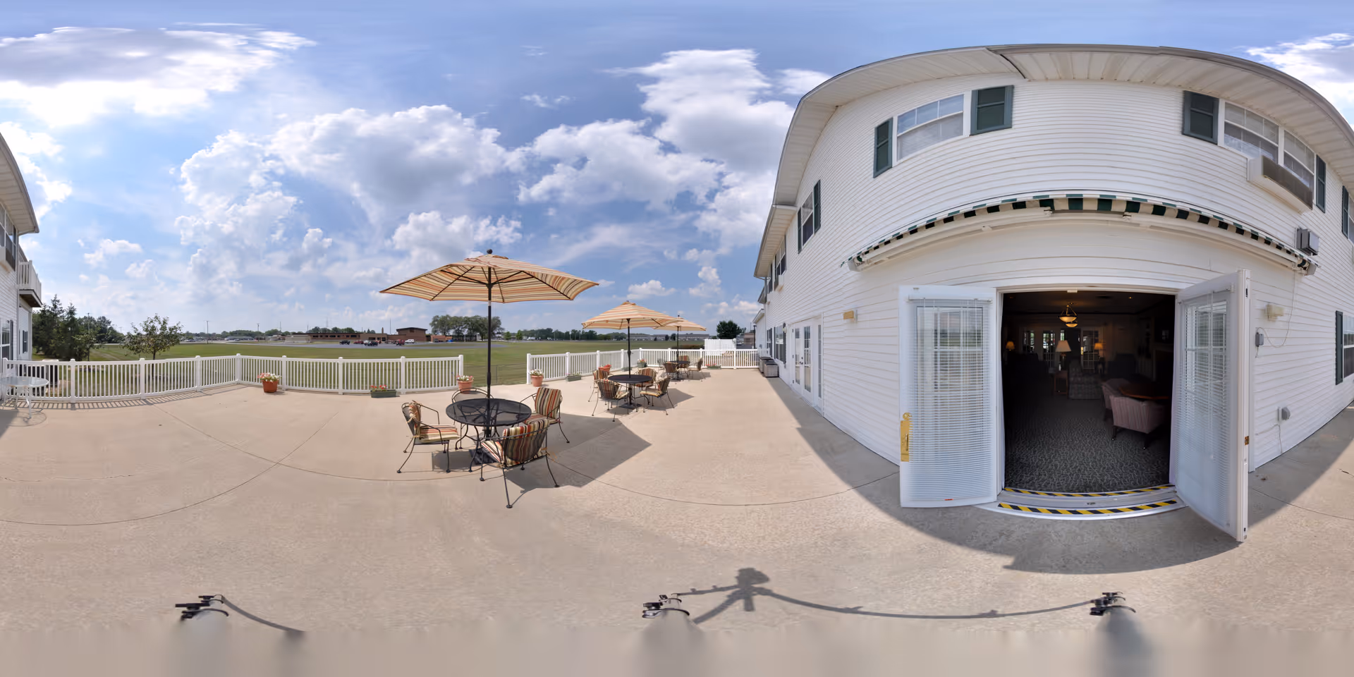 Outdoor patio area at Jefferson Manor with several round tables and chairs under large umbrellas. The patio is spacious with a concrete floor and white railing, overlooking a grassy field under a partly cloudy sky. The building exterior is white with green shutters, and open double doors lead inside to a common area.