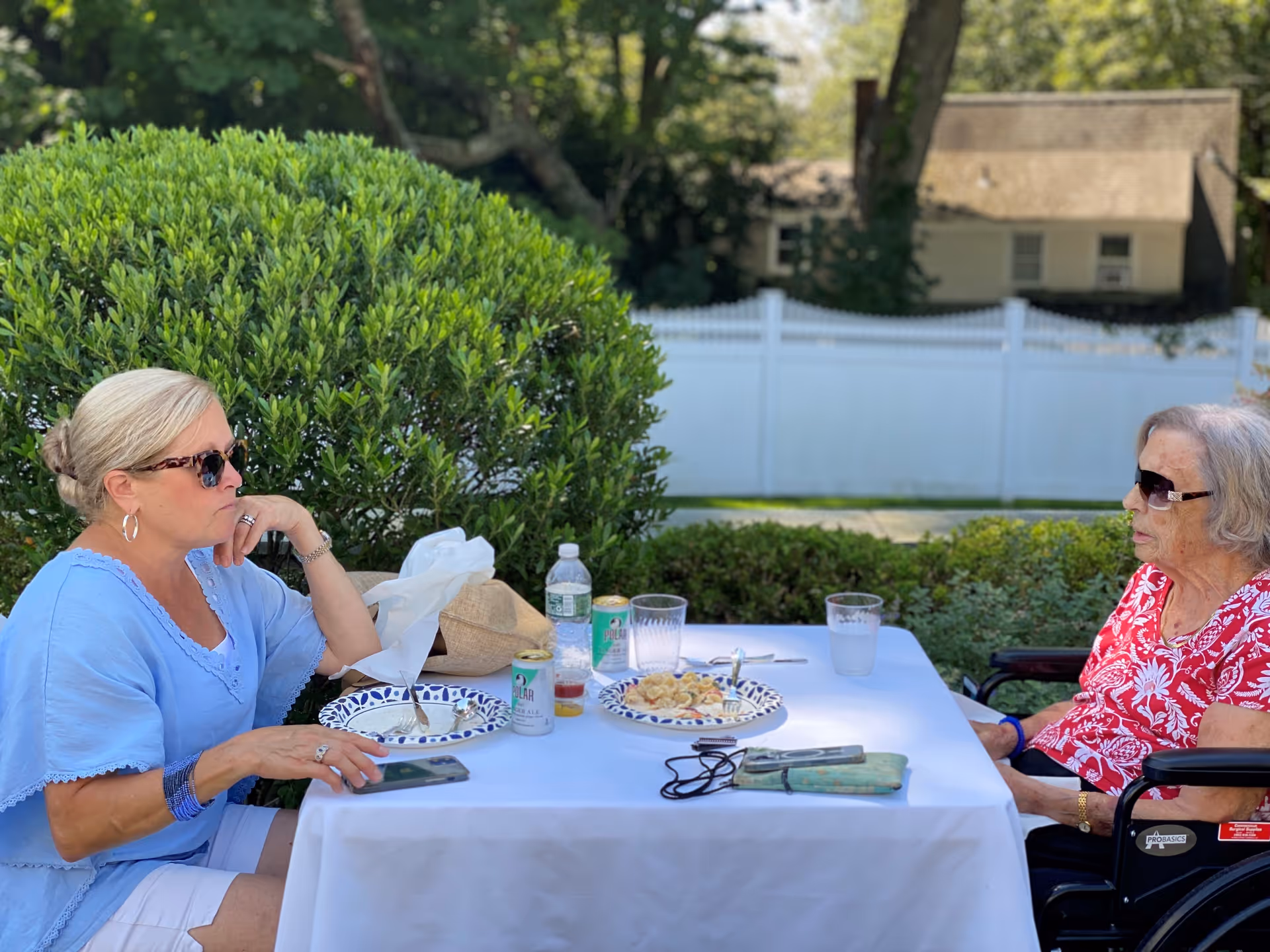 Two women sitting at a white table outdoors having a meal. One woman with blonde hair wearing sunglasses and a light blue top is seated on the left, and an elderly woman with gray hair wearing sunglasses and a red floral top is seated in a wheelchair on the right. There are plates with food, drinks, and utensils on the table, with greenery and a white fence in the background.