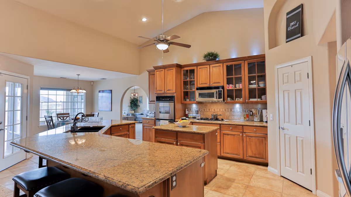 A spacious kitchen with wooden cabinets, granite countertops, a central island, and modern appliances including a microwave and oven. There is a ceiling fan with a light fixture, a dining area with a table and chairs near large windows, and an open archway leading to another room.