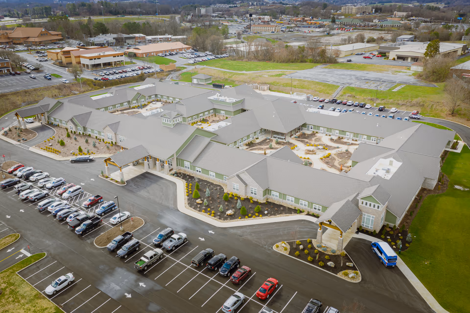 Aerial view of Princeton Transitional Care facility showing a large, single-story building with multiple wings surrounding landscaped courtyards. The building has a gray roof and beige walls with green accents. There is a spacious parking lot with many cars parked, and surrounding areas include grassy fields and other buildings in the distance.