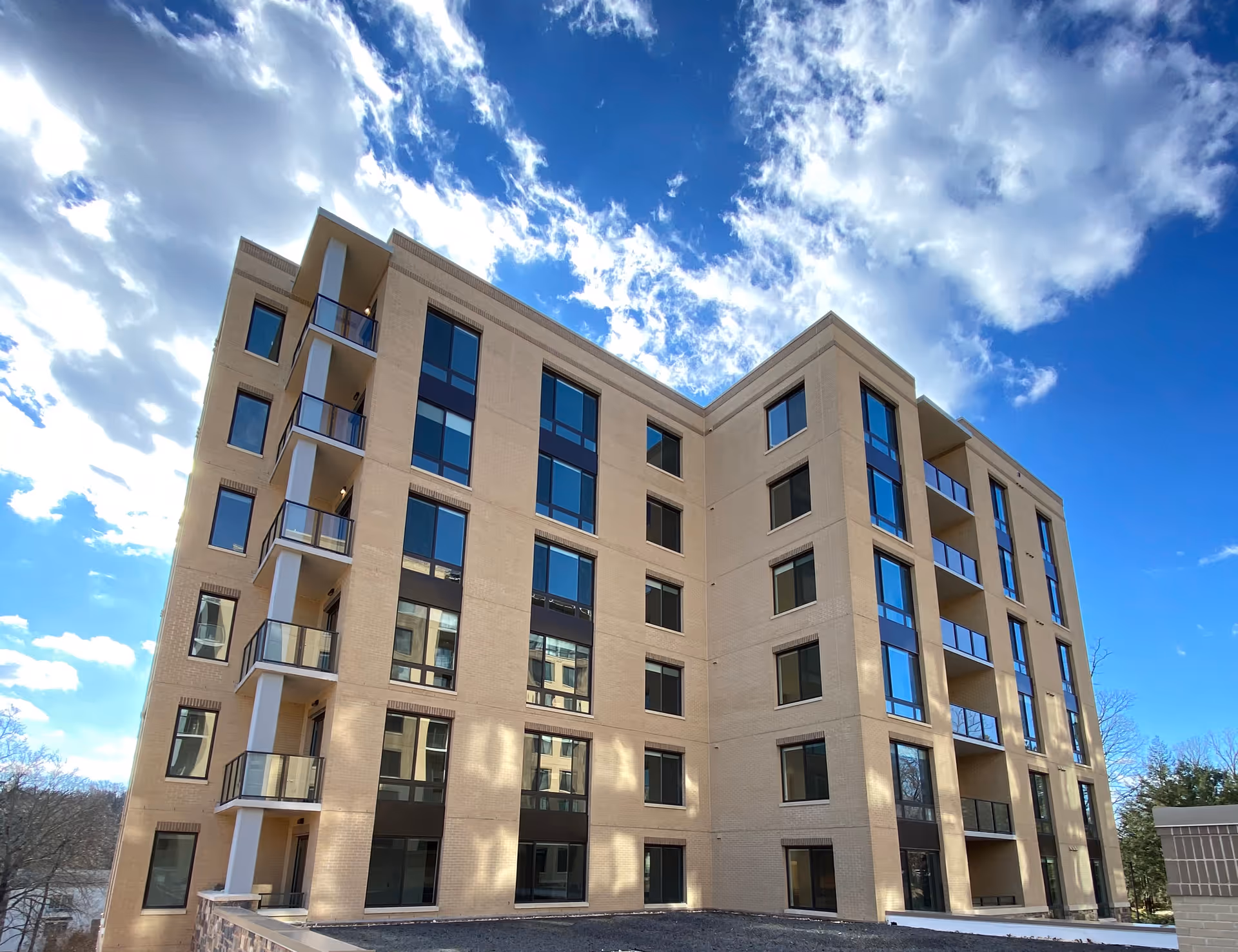 Exterior view of a modern multi-story beige brick building with large windows and balconies under a partly cloudy blue sky.