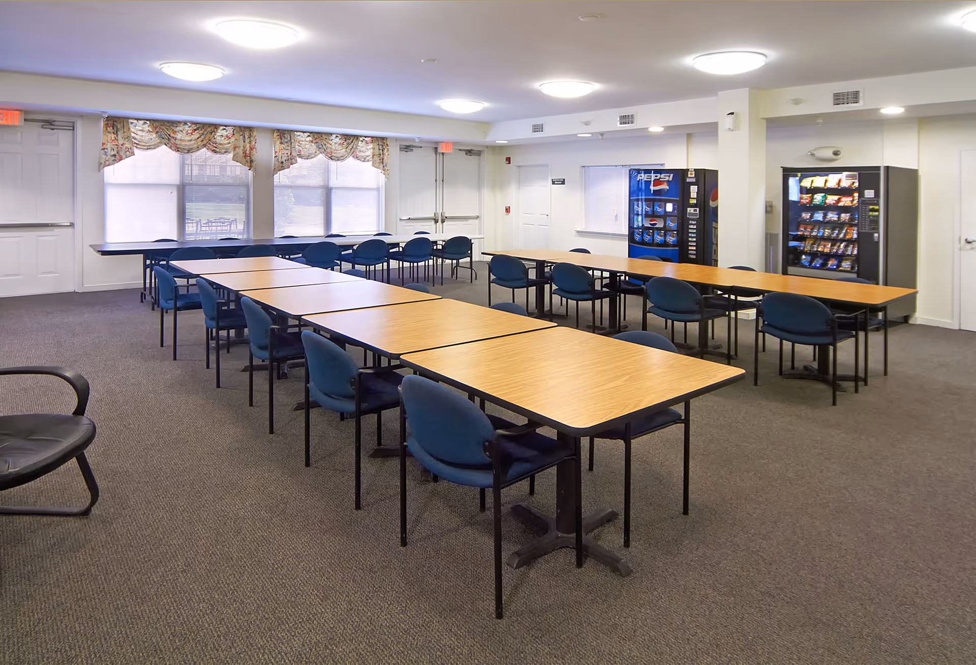 Large multipurpose dining room with rows of tables and blue chairs and vending machines along the back wall.