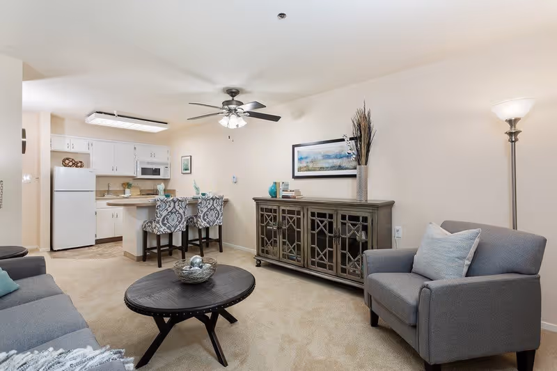 A cozy living room area with a gray armchair and sofa, a round black coffee table with decorative items, a wooden cabinet with glass doors, and a floor lamp. In the background, there is a kitchen with white cabinets, a refrigerator, microwave, and a small island with two patterned bar stools. The room has beige carpet and walls, with a ceiling fan and a framed picture on the wall above the cabinet.