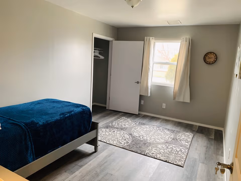 A simple bedroom with a single bed covered in a dark blue blanket, a patterned area rug on the floor, a window with white curtains, a wall clock, and an open closet door showing hangers inside.