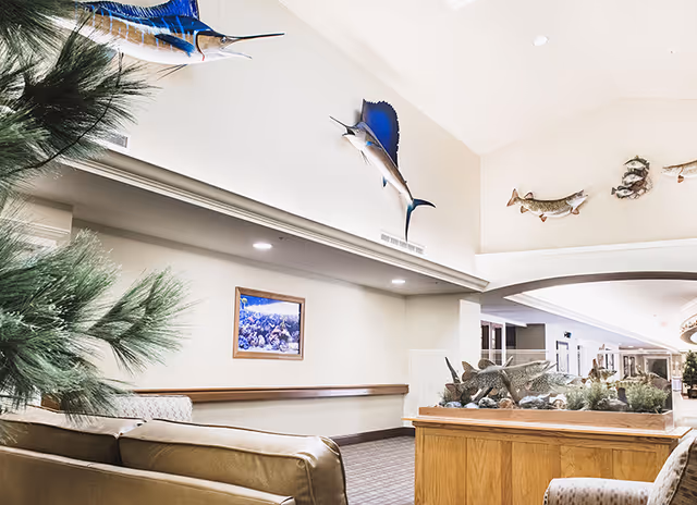 Interior view of a senior living facility lounge area with beige sofas, a large fish tank on a wooden stand, and mounted fish decorations on the walls. There is a framed underwater-themed picture on the wall and some greenery in the foreground.