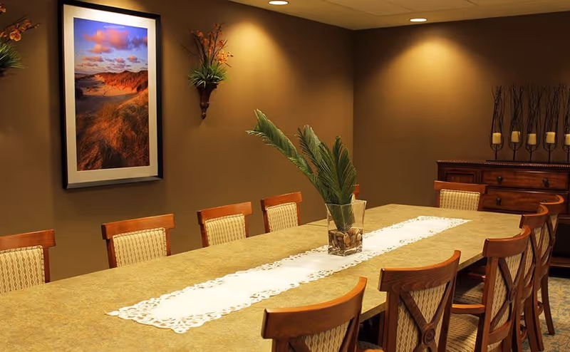 A dining room with a long rectangular table surrounded by wooden chairs with cushioned seats. The table has a white lace runner and a glass vase with green leaves and stones. The walls are painted brown and decorated with a framed landscape photograph, a wall-mounted plant, and a wooden sideboard with candle holders.
