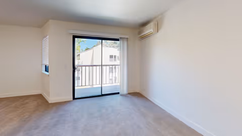 Empty carpeted living room with a sliding glass door to a balcony, a small side window, and a wall-mounted air conditioning unit.