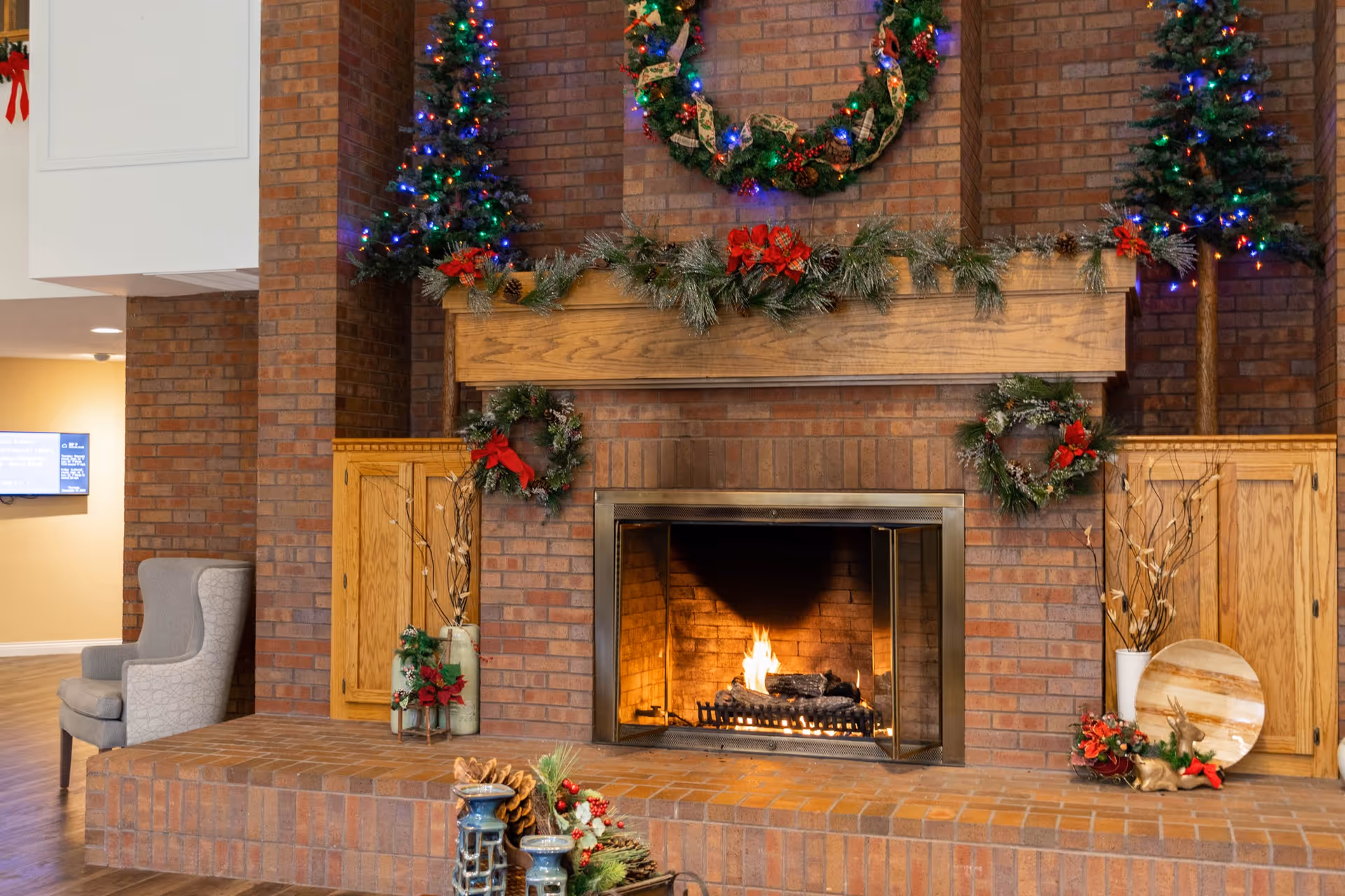 A cozy brick fireplace decorated for the holidays with a wreath above the mantel, garlands, two small Christmas trees with colorful lights on either side, and additional holiday decorations including pine cones and red bows. A gray armchair is visible to the left, and wooden cabinets flank the fireplace.