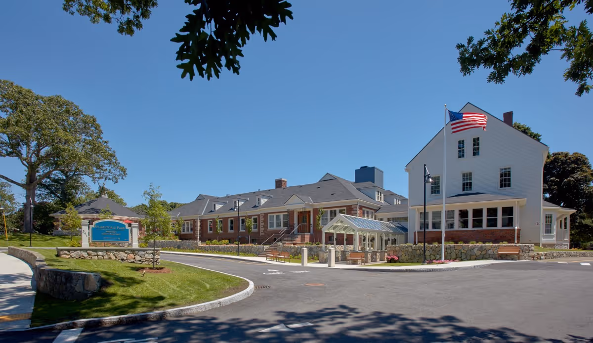 Front exterior of a senior living facility with a circular driveway, landscaped lawn, entrance canopy, and an American flag.