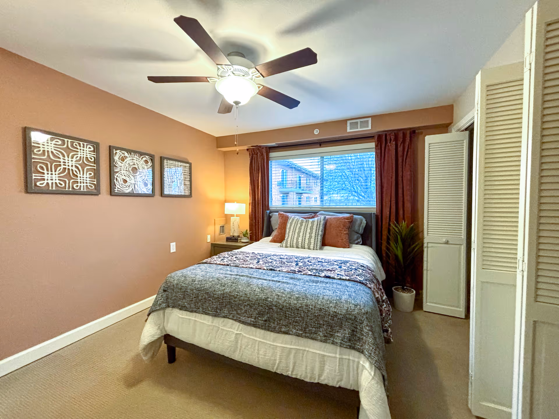 Neat bedroom with a bed, pillows and throw, ceiling fan, window with curtains, nightstand and wall art.
