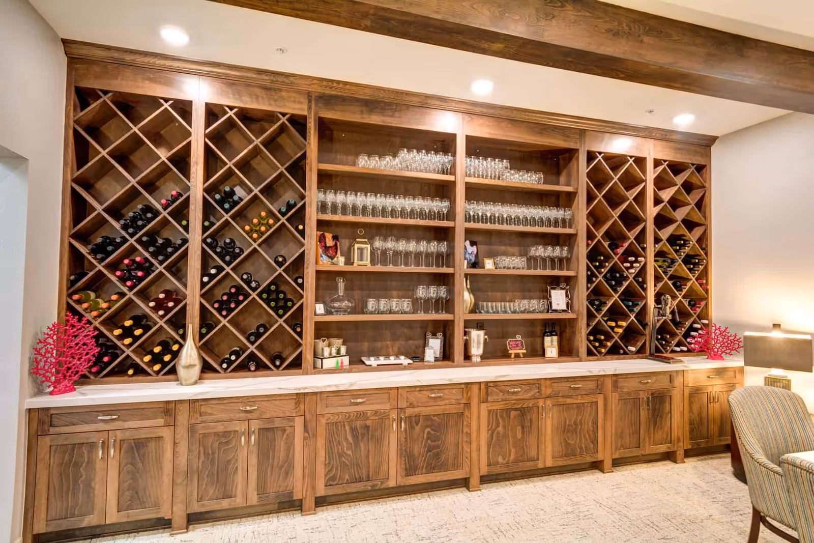 A wooden wine rack and shelving unit filled with wine bottles and various types of glassware, including wine glasses and tumblers, in a well-lit room with a lamp and a striped upholstered chair partially visible on the right.