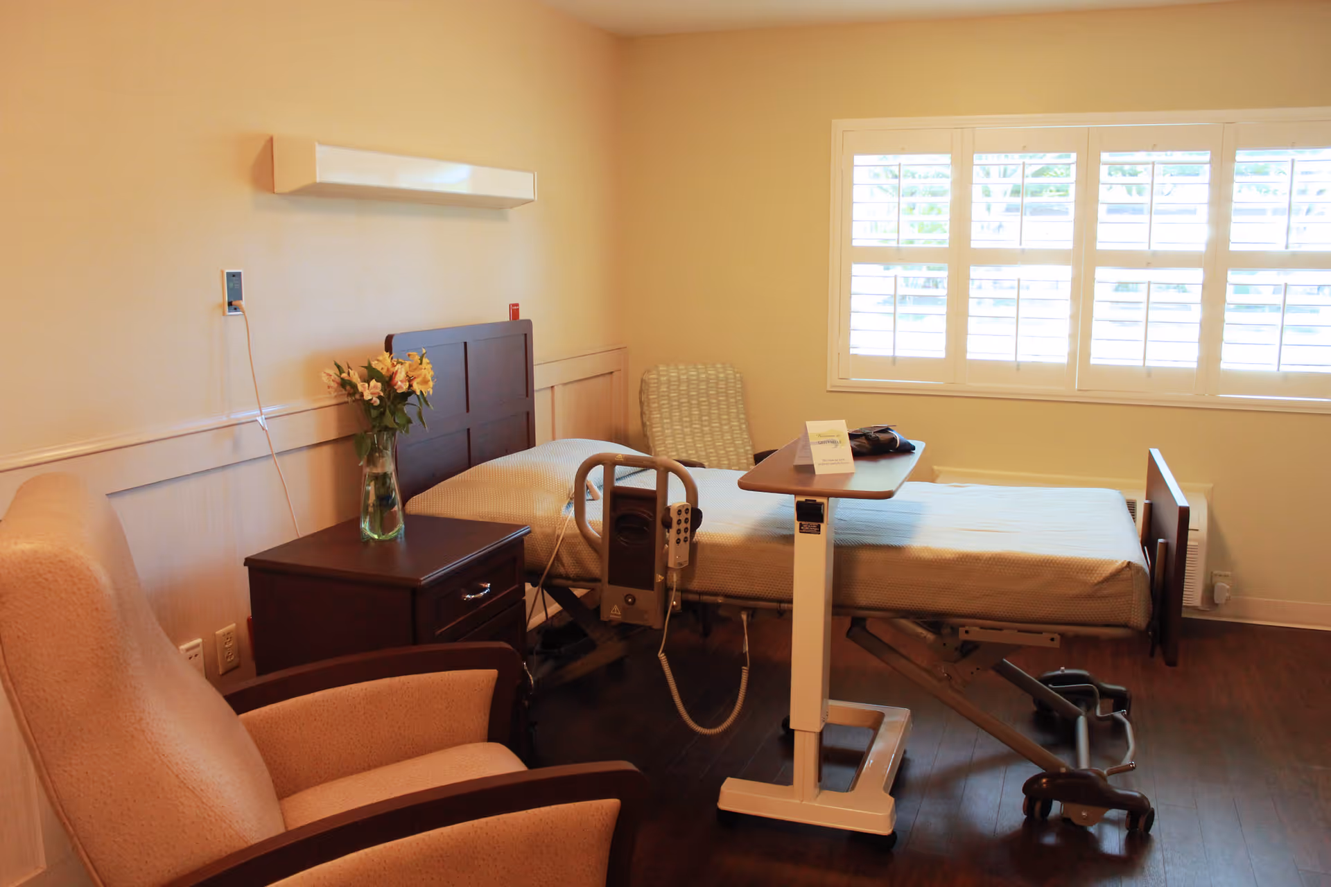 A well-lit room with a hospital-style adjustable bed, a small overbed table, a wooden nightstand with a vase of flowers, a cushioned armchair, and a window with white shutters allowing natural light to enter.