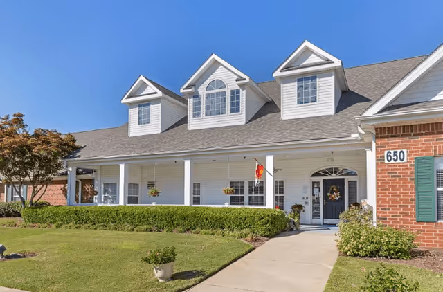 Front exterior view of a single-story building with white siding and a gray shingled roof featuring three dormer windows. The building has a covered porch with white columns, hanging flower baskets, and a flag. There is a well-maintained lawn and shrubs in front, with a concrete walkway leading to a dark-colored front door decorated with a wreath. The building number 650 is visible on a brick section to the right.