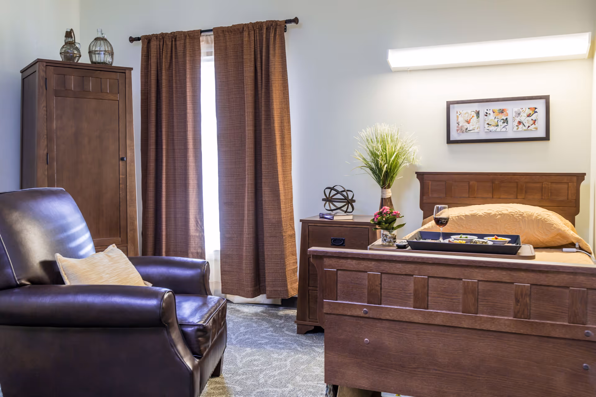 A cozy bedroom in a senior living facility featuring a wooden bed with a beige bedspread, a tray with a meal and a glass of wine on the bed, a wooden nightstand with decorative items and a vase with green plants, a dark brown leather armchair with a beige pillow, a wooden wardrobe, and brown curtains covering a window.