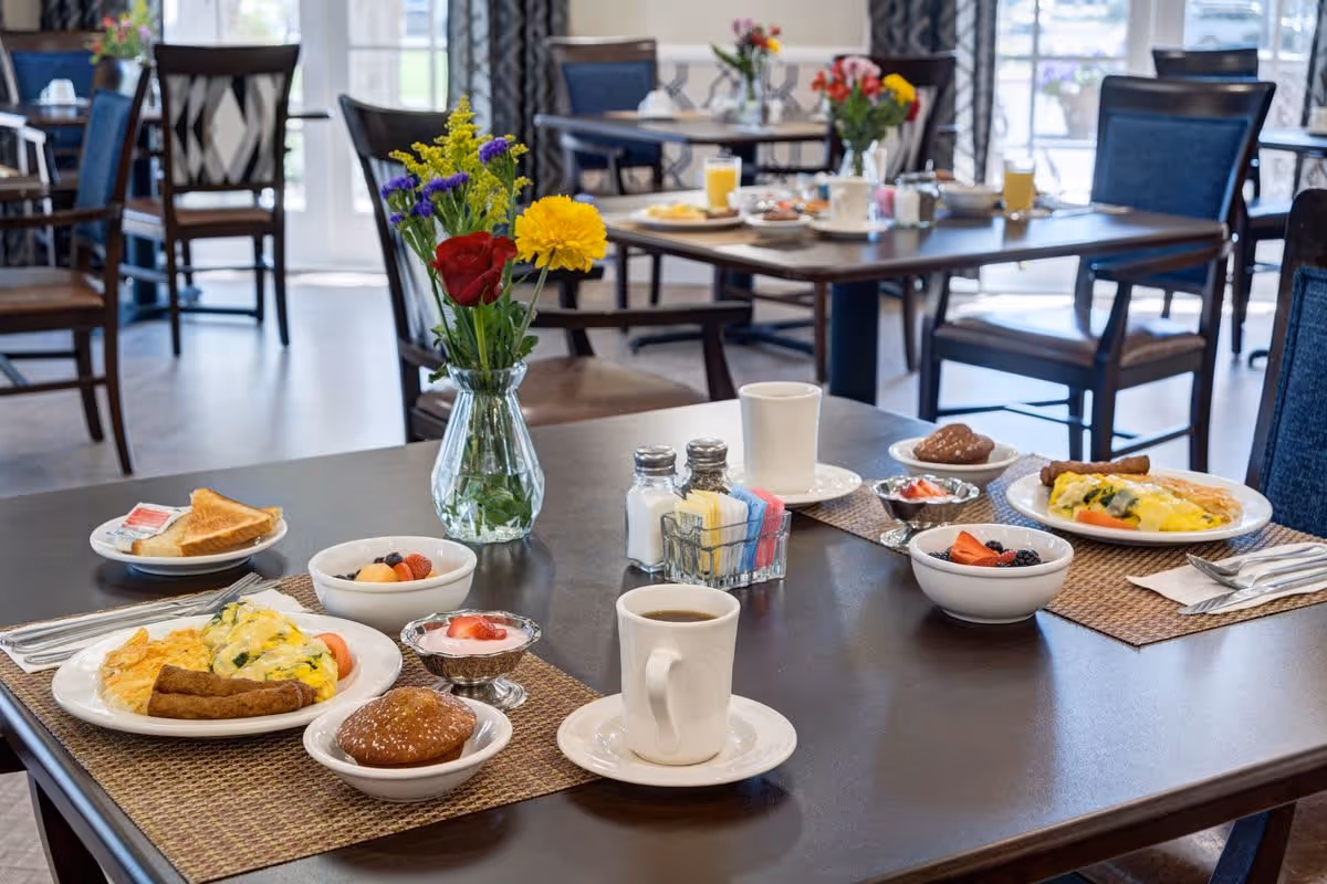 A dining table set for breakfast with plates of scrambled eggs, sausage, toast, fruit bowls, muffins, coffee cups, and a vase with colorful flowers in a bright dining room with multiple tables and chairs.