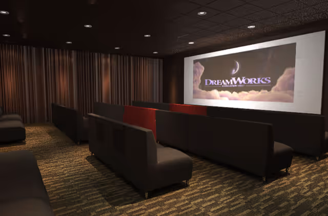 A small movie theater room with rows of dark cushioned benches facing a large screen displaying the DreamWorks logo. The room has dim lighting, carpeted floor, and dark curtains along the back wall.