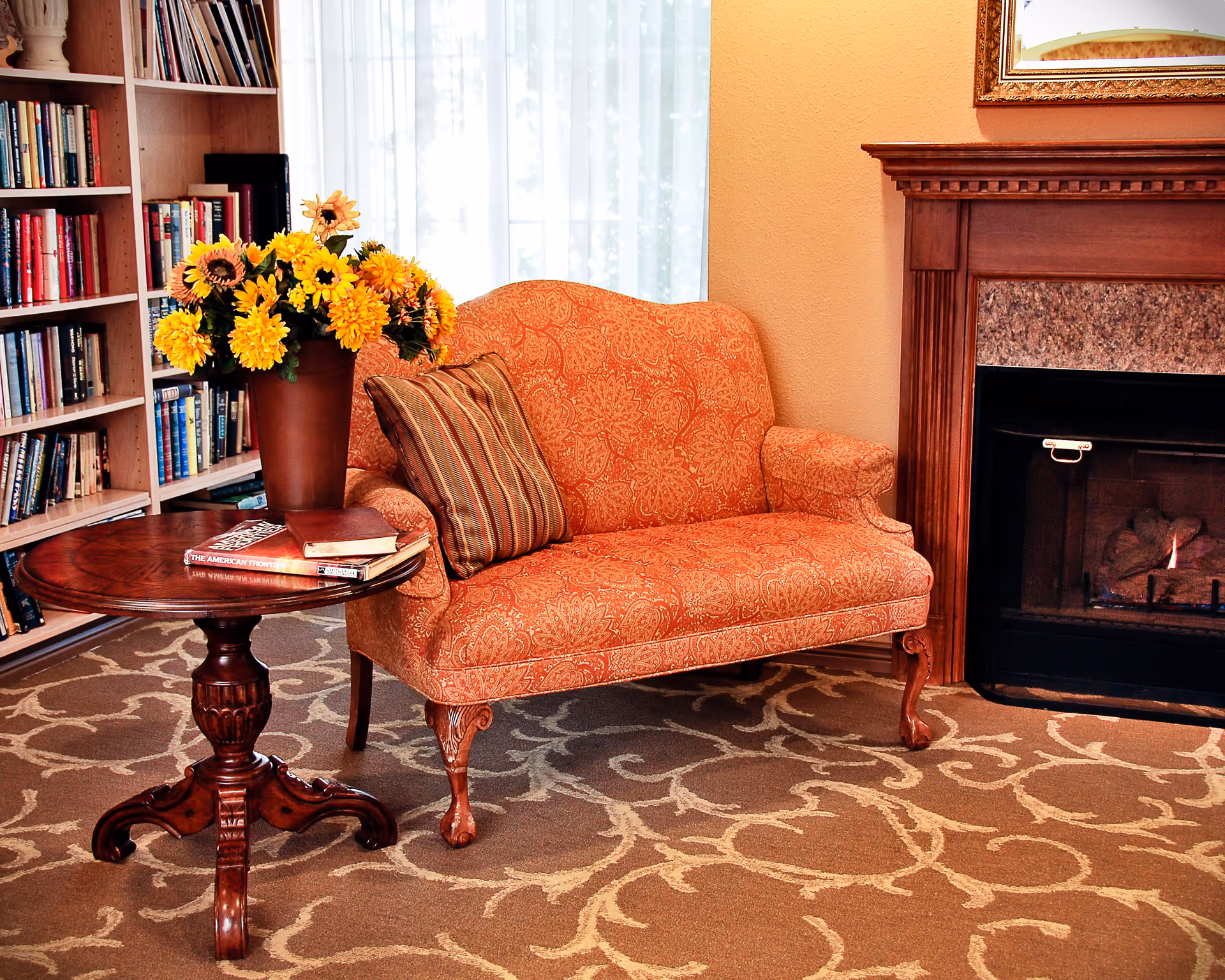 A cozy living room area featuring an orange patterned loveseat with a striped pillow, a wooden round table with a vase of yellow flowers and several books on it, a bookshelf filled with books, and a fireplace with a wooden mantle and a fire burning inside.