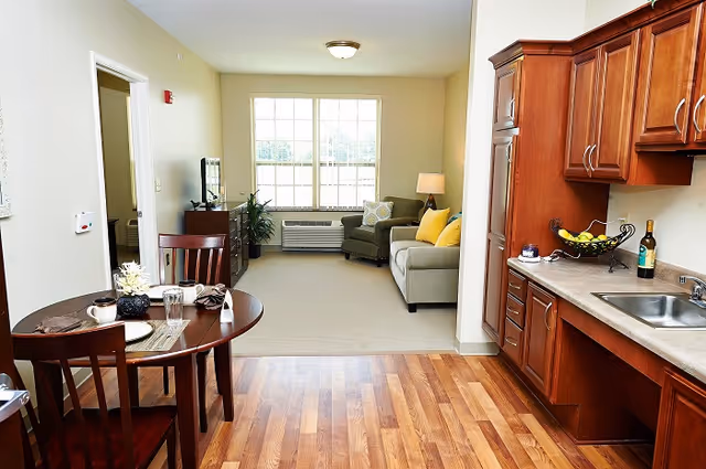 Open-plan senior apartment with a small kitchen and dining table leading to a bright living area with a sofa and large window.