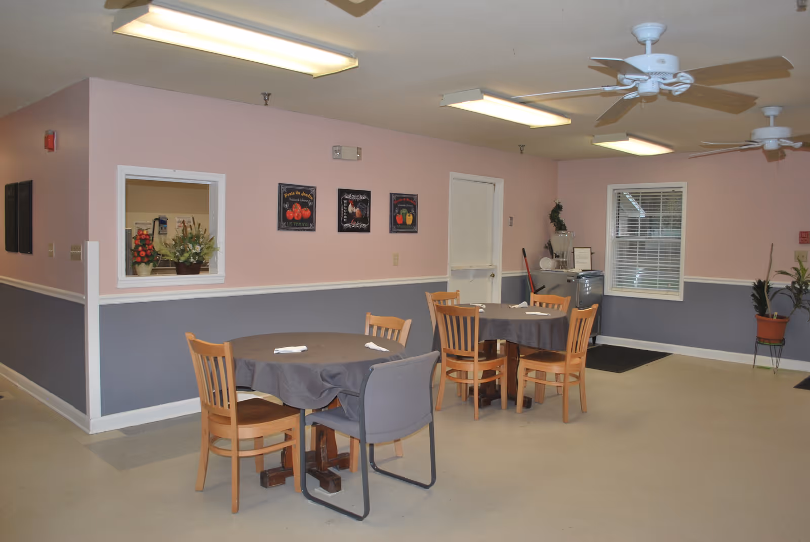 Interior view of a dining area in an assisted living facility with two round tables covered with gray tablecloths, each surrounded by wooden chairs and one gray cushioned chair. The walls are painted pink on the upper half and gray on the lower half, decorated with three framed pictures. There are ceiling fans and fluorescent lights overhead, a window with blinds, a small refrigerator with a water dispenser, and some potted plants in the corner.