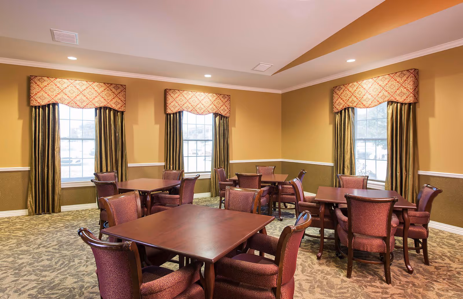 A dining room with several square wooden tables surrounded by upholstered chairs. The room has beige walls with a white chair rail and three large windows with patterned valances and dark curtains. The carpet has a floral pattern and recessed ceiling lights illuminate the space.