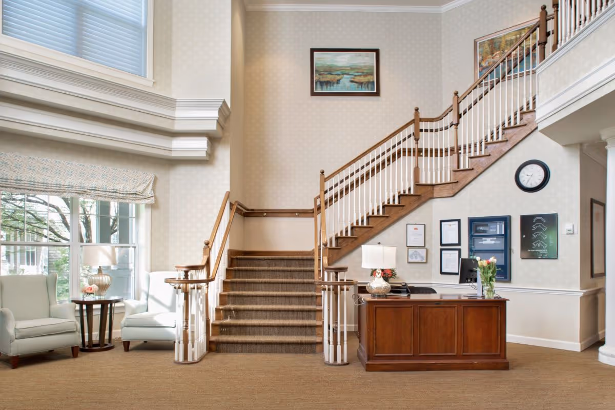 Interior view of a senior living facility lobby with a wooden reception desk on the right, a staircase with wooden handrails leading to the upper floor in the center, two light-colored armchairs with a small round table between them near large windows on the left, and framed artwork and a clock on the walls.