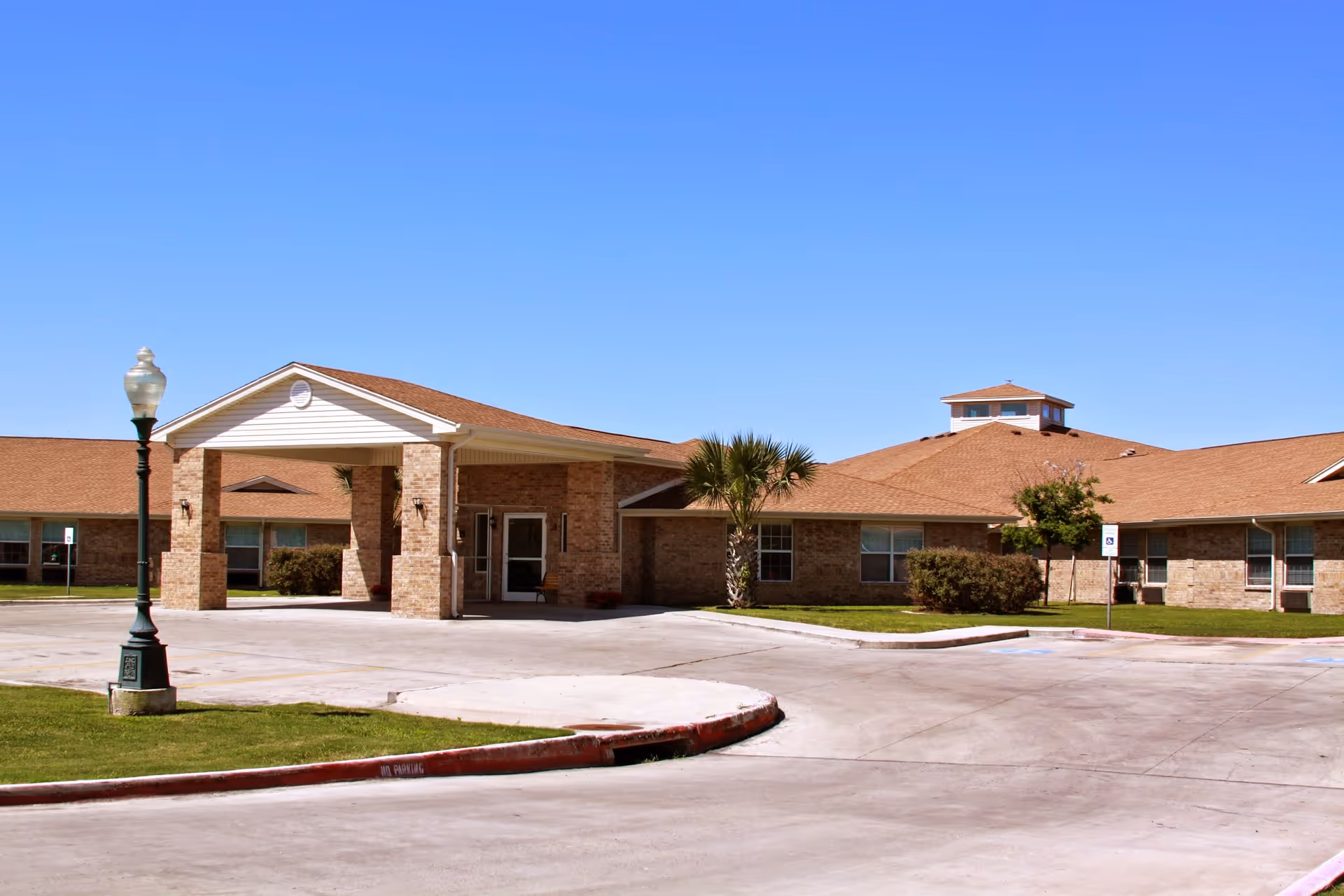 Exterior view of Corpus Christi Nursing and Rehabilitation Center showing a single-story brick building with a covered entrance, a lamp post, palm trees, and a clear blue sky.