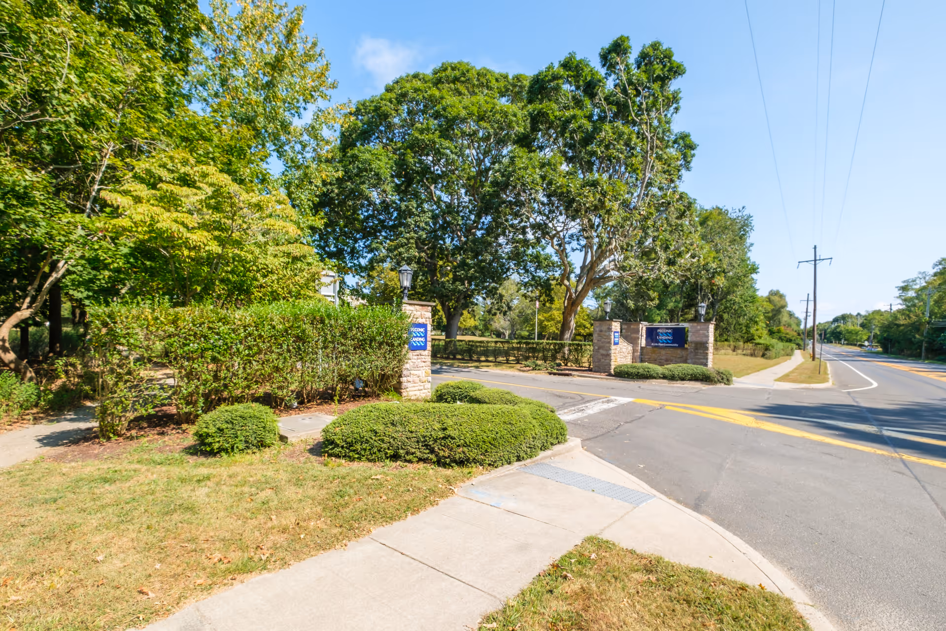 Entrance to Peconic Landing facility with stone pillars and signs, surrounded by green bushes and trees under a clear blue sky, next to a paved road and sidewalk.