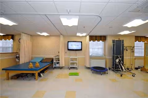 Rehab room with therapy tables, curtains, a wall TV, and exercise equipment.