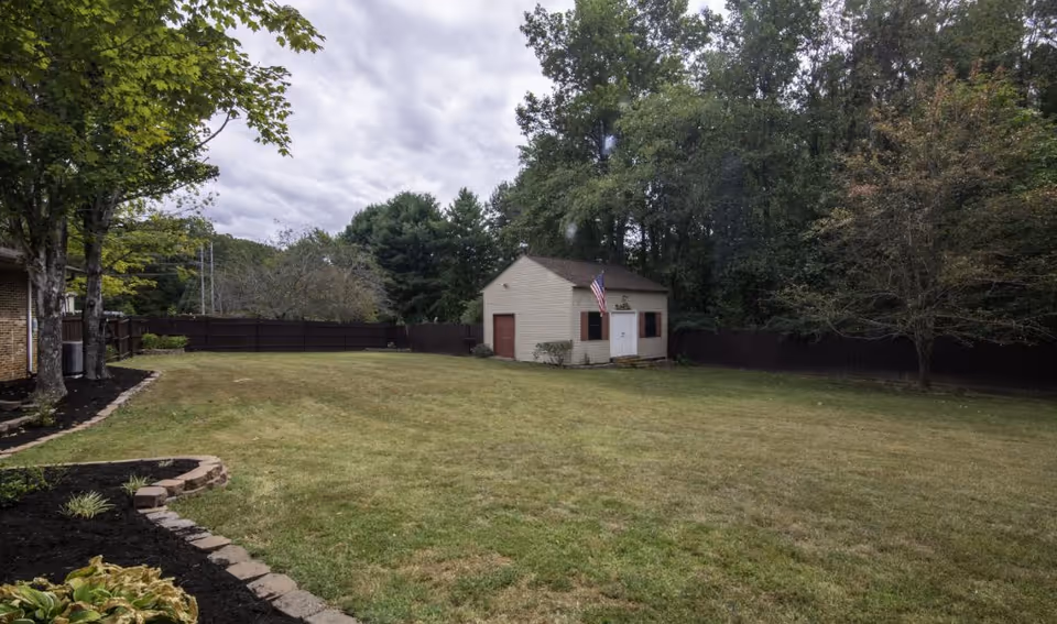 A large grassy backyard area with a small beige shed featuring a red door and white double doors, an American flag mounted on the shed, surrounded by a dark wooden fence and trees under a cloudy sky.