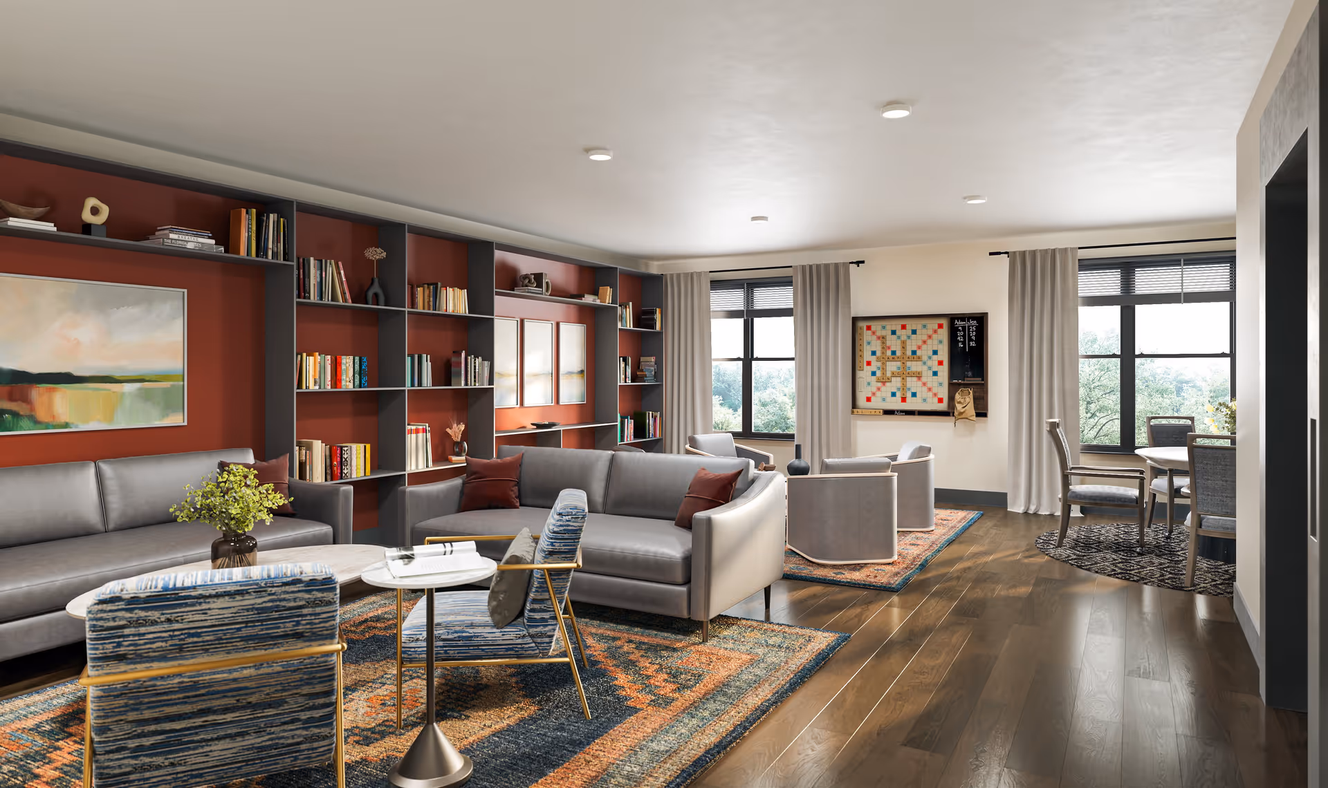 A spacious and well-lit living room area with modern furniture including two gray sofas, two blue patterned armchairs, and a small round table. The room features a large built-in bookshelf with books and decorative items against a red accent wall. There are two large windows with beige curtains letting in natural light. A Scrabble game board is mounted on the wall near a small seating area with four gray chairs arranged around a rug. Hardwood floors and colorful area rugs add warmth to the space.