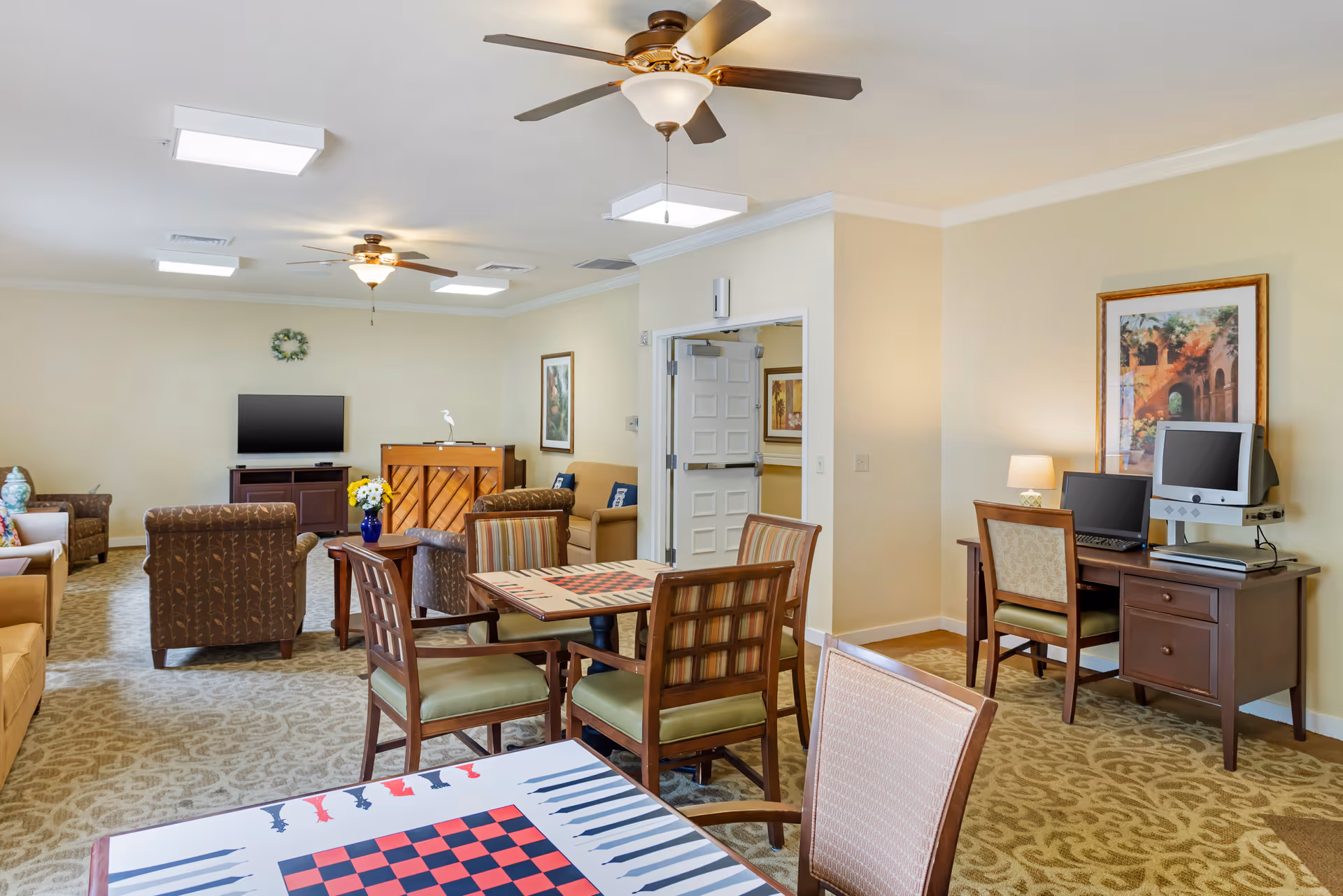 A spacious senior living common area with multiple seating arrangements including armchairs and sofas. There are two tables with checkerboard and backgammon game designs, a piano, a flat-screen TV on a stand, and a desk with two computers. The room has beige walls, patterned carpet, ceiling fans, and framed artwork on the walls.