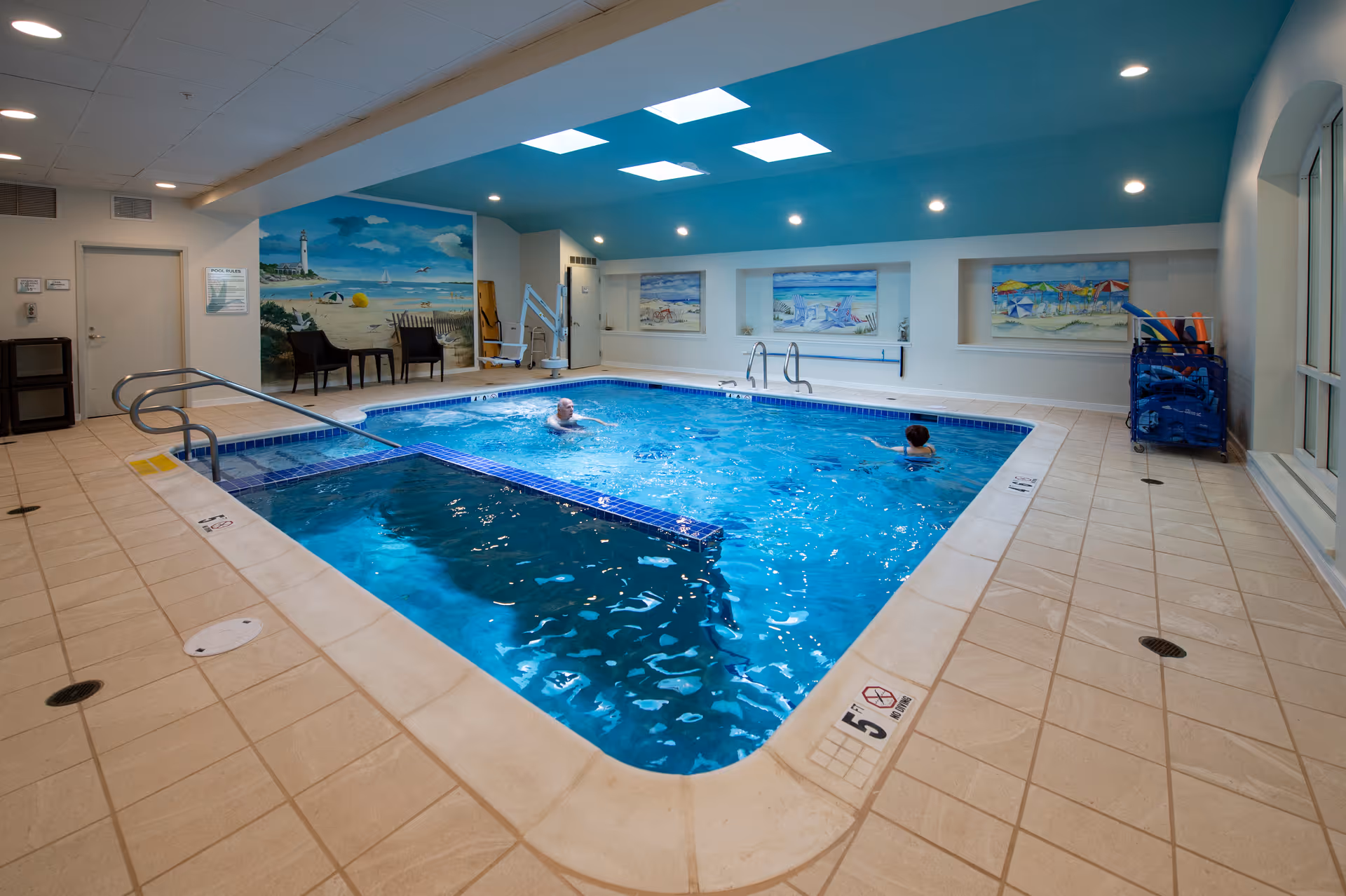 Indoor swimming pool with two people swimming. The pool area has beige tiled flooring, a blue ceiling with skylights, and beach-themed murals on the walls. There are chairs and pool equipment along the sides.