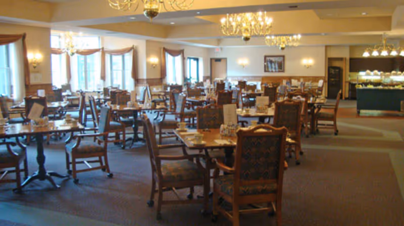 A spacious dining room with multiple wooden tables and chairs arranged neatly. Each table has cups, saucers, and menus placed on it. The room is well-lit with large windows allowing natural light and chandeliers hanging from the ceiling. There is a buffet area visible in the background.