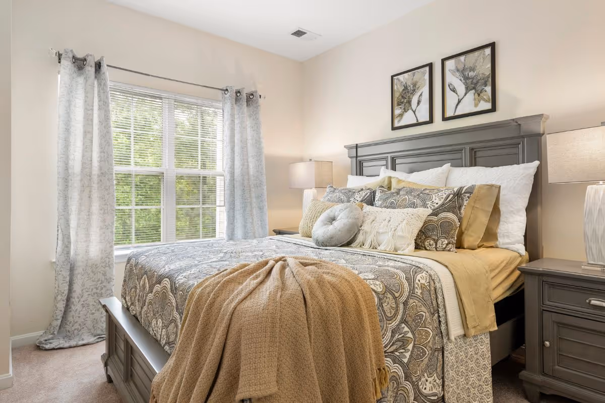 Well-decorated bedroom with a large bed dressed in patterned bedding, multiple pillows, nightstands with lamps, and a window with curtains.