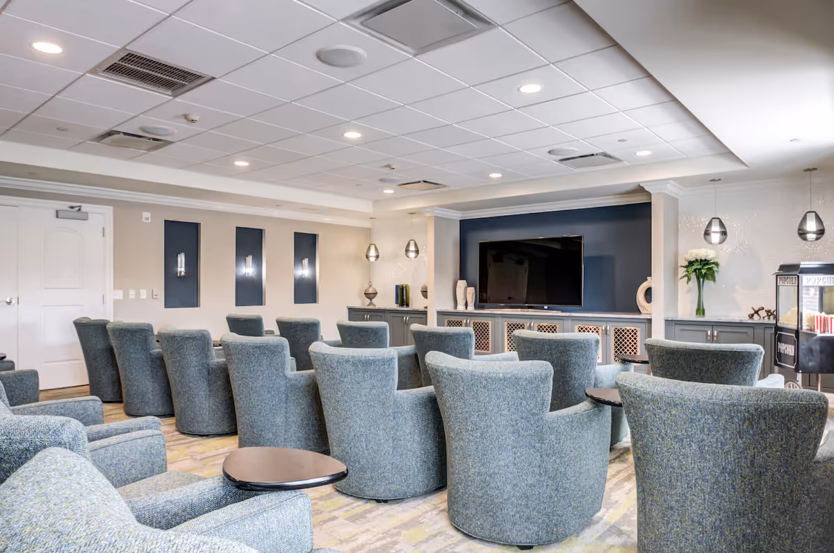 A modern common area with multiple blue upholstered armchairs arranged in rows facing a large flat-screen TV mounted on a dark accent wall. The room features a light-colored ceiling with recessed lighting, decorative wall sconces, and pendant lights. There is a popcorn machine on the right side and cabinets with decorative items underneath the TV.