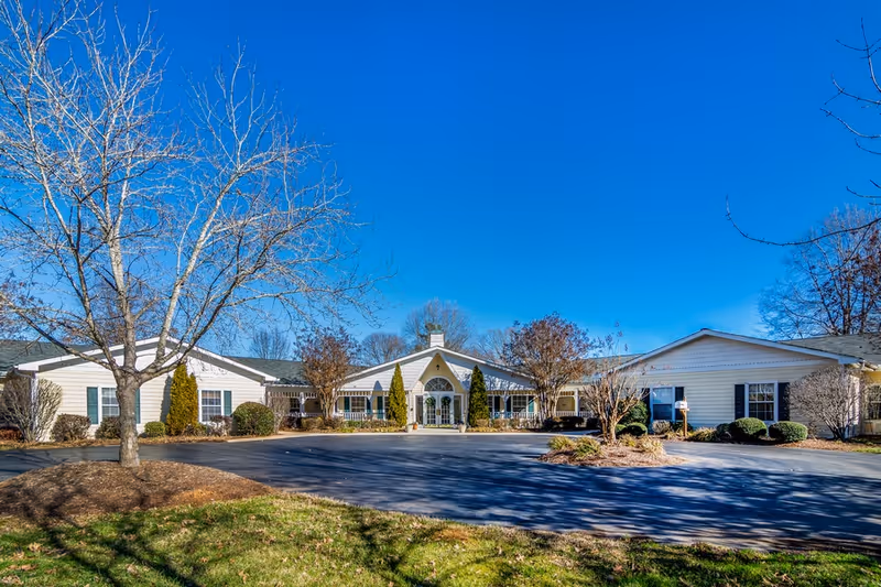 Exterior view of a single-story senior living facility building with white siding, a central entrance with large windows, surrounded by leafless trees and shrubs under a clear blue sky.