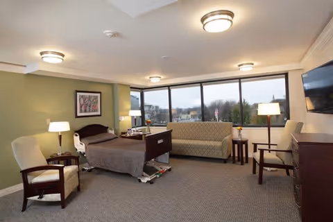 A spacious senior living room with a hospital-style bed, two armchairs, a sofa, a flat-screen TV mounted on the wall, and large windows offering a view of trees and buildings outside. The room is well-lit with ceiling lights and floor lamps, and has a carpeted floor and light green walls.