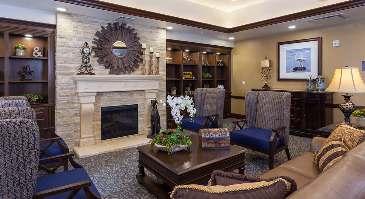 A cozy senior living room featuring a stone fireplace with a decorative round mirror above it, surrounded by built-in wooden shelves with various decor items. The room has comfortable armchairs with blue cushions, a brown leather sofa with patterned pillows, a wooden coffee table with a floral arrangement and a decorative chest, and warm lighting from table lamps.