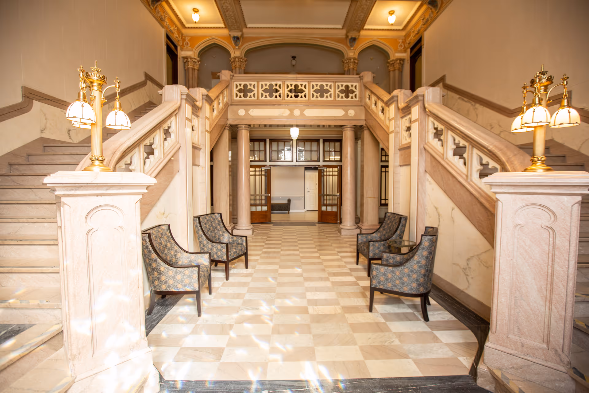 Interior view of a senior apartment building lobby featuring a grand marble staircase with ornate railings on both sides. There are six patterned armchairs arranged in two groups of three along the walls. The floor is checkered marble, and the ceiling has decorative molding with warm lighting fixtures.