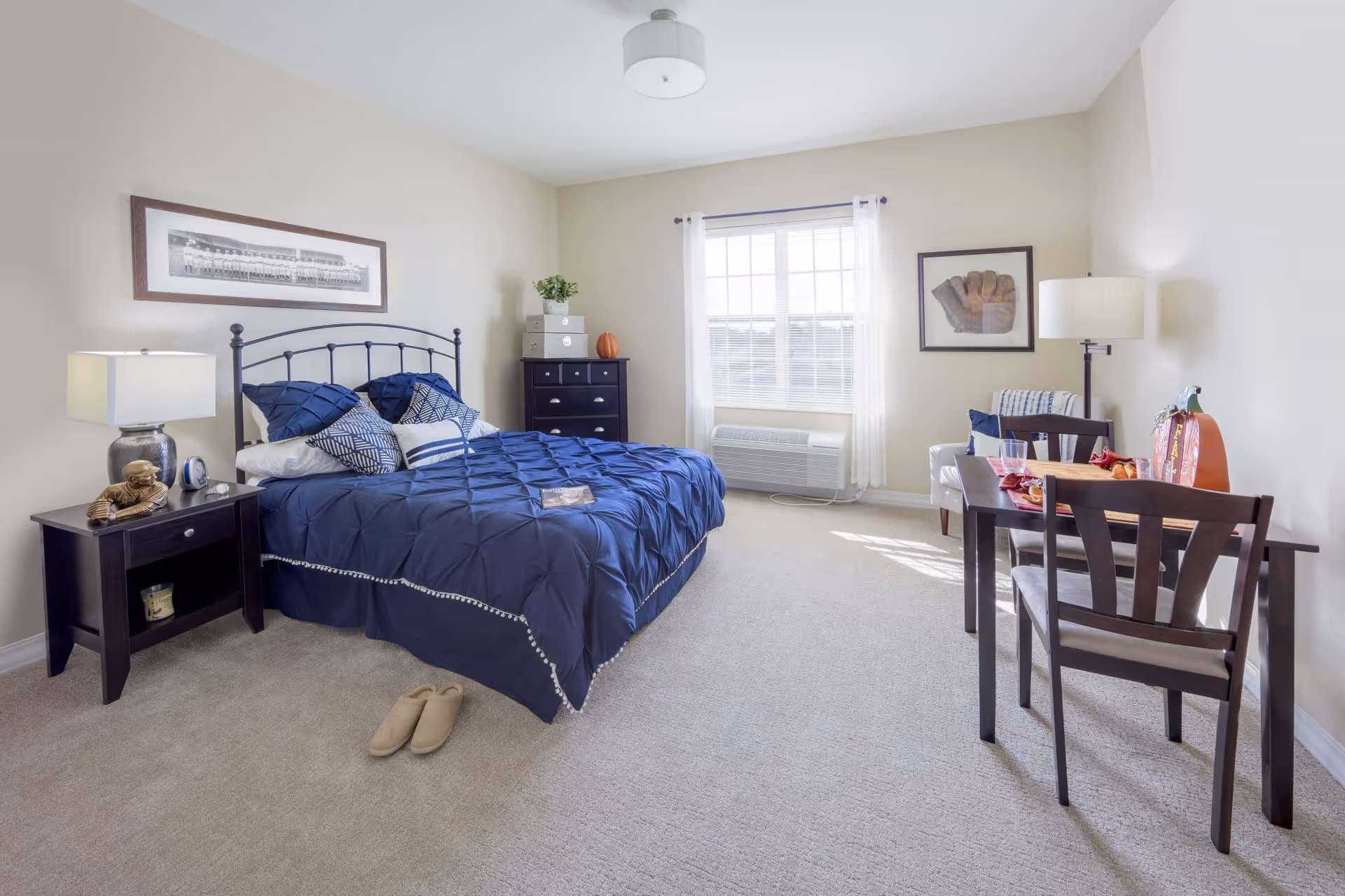 Bright furnished bedroom with a navy bedspread, nightstands, dresser, window, and a small dining table and chairs.