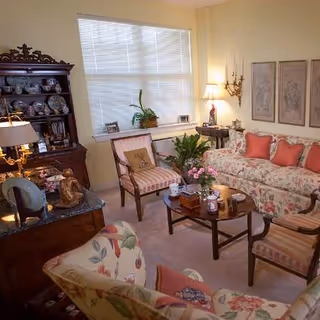 Cozy living room with floral-upholstered sofa and chairs, a wooden china cabinet, coffee table, lamps, and decorative plants under a large window.