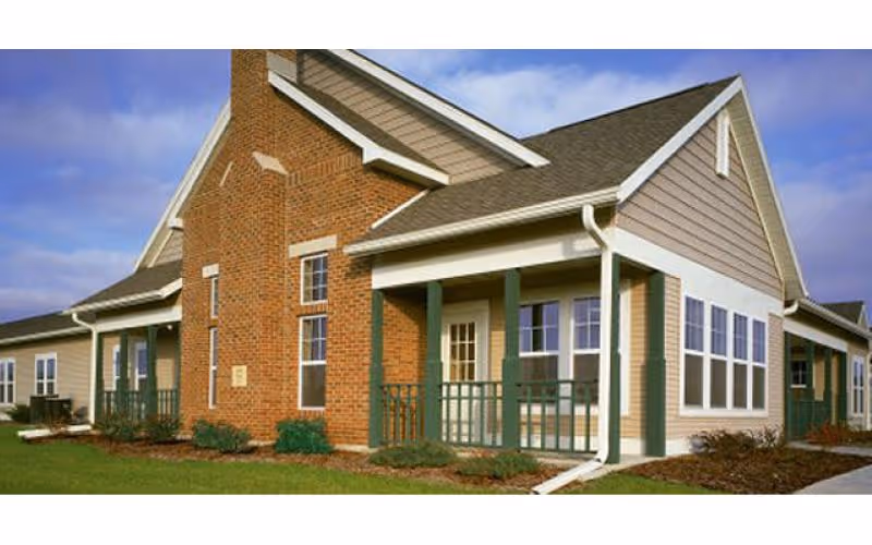 Exterior view of a single-story senior living facility building with a combination of brick and beige siding, green porch railings, multiple windows, and a well-maintained lawn under a partly cloudy sky.