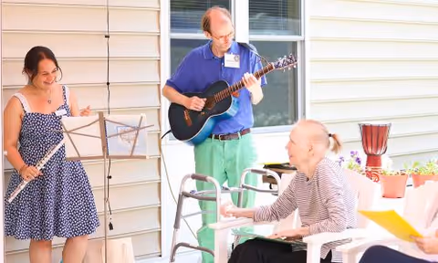 A woman playing the flute and a man playing the guitar entertain an elderly woman seated outdoors near a white building with windows. The elderly woman is smiling and reaching out her hand, and there are potted plants on a table nearby.