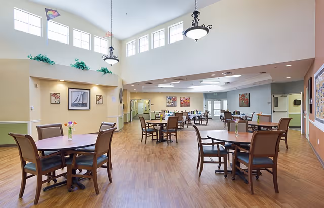 A spacious and well-lit dining area with multiple round wooden tables and chairs arranged neatly. The room features high ceilings with large windows near the top, allowing natural light to fill the space. There are decorative paintings on the walls and small flower vases on each table. The flooring is wood, and the walls are painted in soft, warm colors.