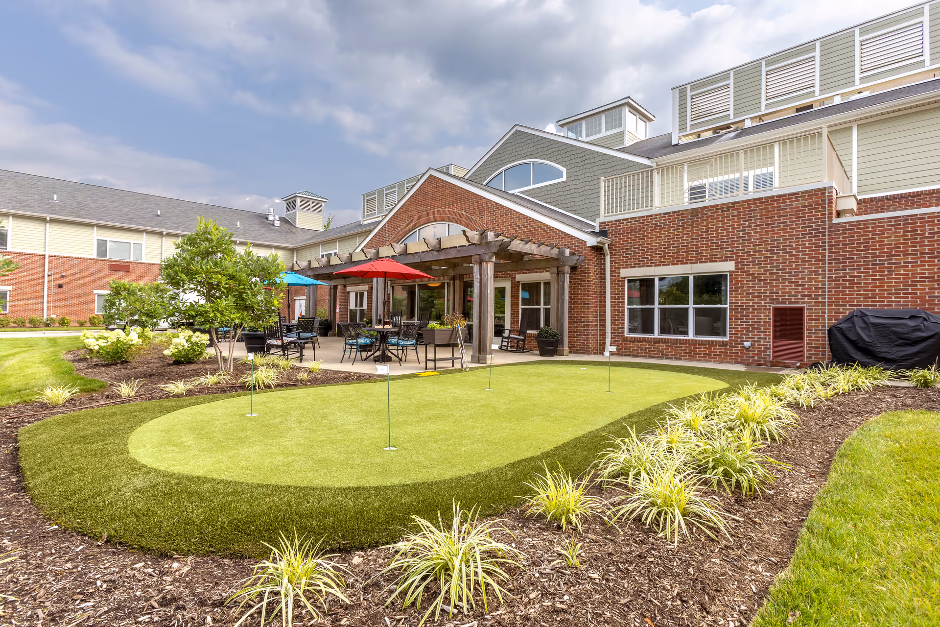 Outdoor area of a senior living facility featuring a putting green surrounded by landscaped plants and mulch. In the background, there is a brick building with a covered patio area furnished with tables, chairs, and colorful umbrellas under a partly cloudy sky.