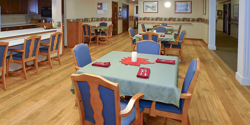 Communal dining room with green-clothed square tables set with napkins and utensils, wooden chairs with blue upholstery, a serving counter with bar stools, and hardwood floors.