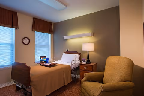 A cozy bedroom in a senior living facility with a single bed covered in a brown blanket, a bedside table with a lamp and a clock on the wall. There is a comfortable armchair next to the bed and two windows with brown valances letting in natural light.